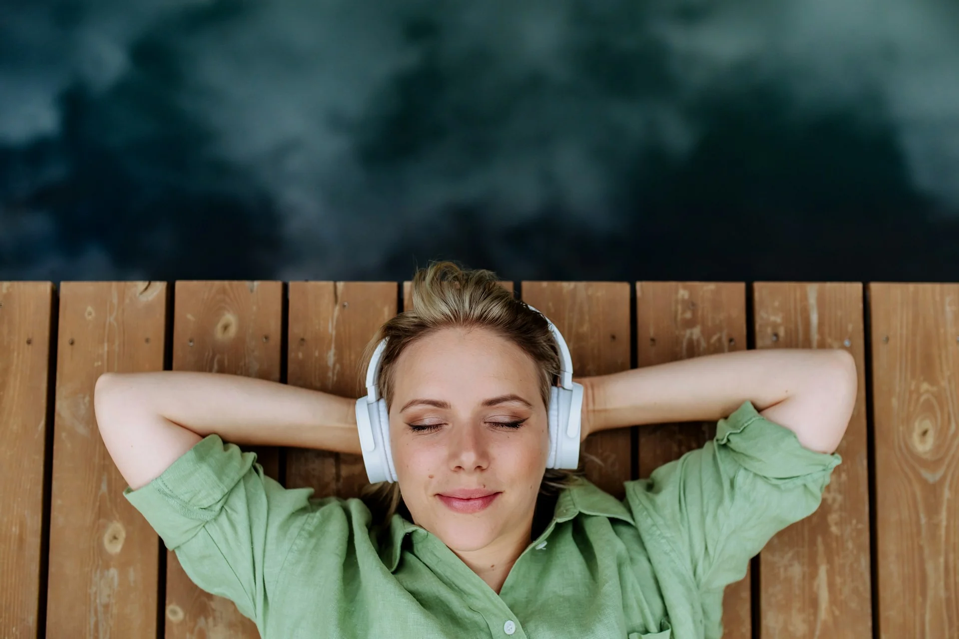 A woman relaxes by a lake with white headphones on, listening to music