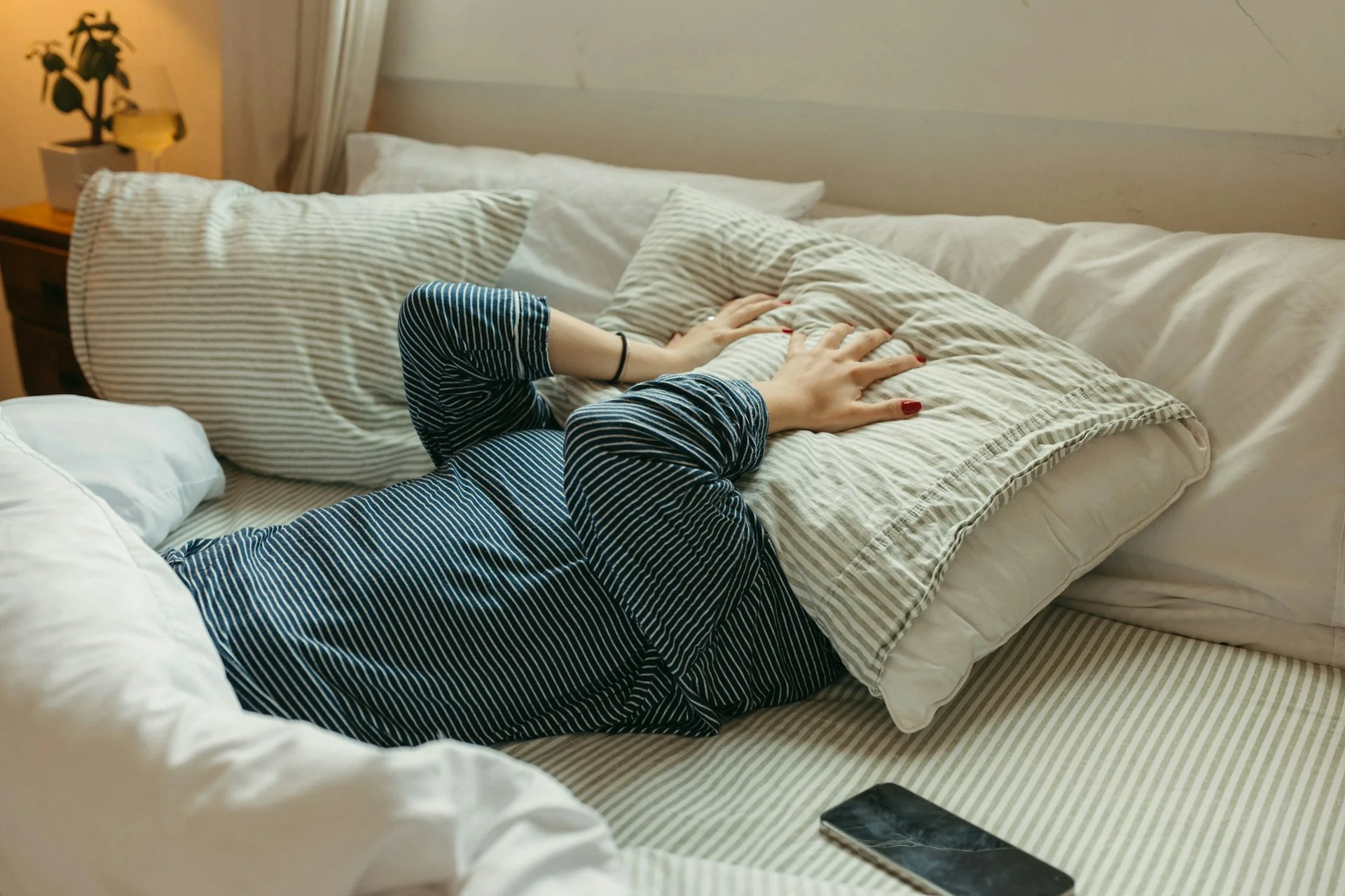 A woman lays in bed with a hangover, covering her face totally with a pillow, as though to block out sound and light