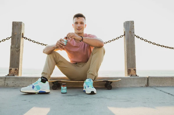 A guy sits on his skateboard on a beach boardwalk holding a bottle of Sunday Scaries Vegan AF gummies