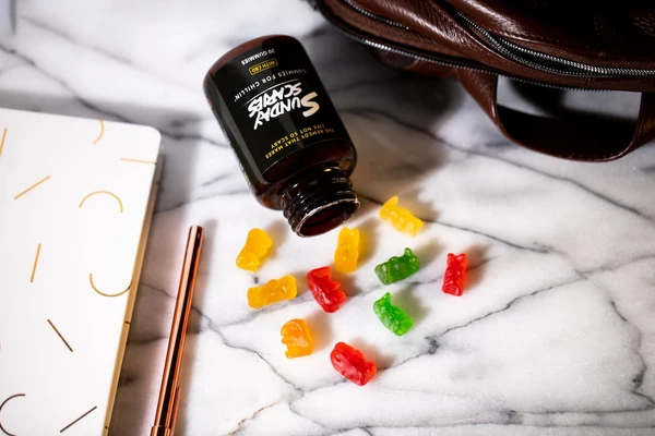 An open bottle of Sunday Scaries CBD gummies pours onto a counter next to a notepad and work bag