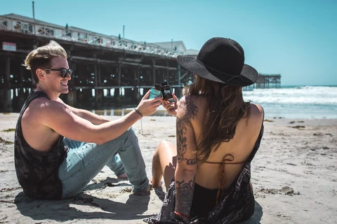 A couple sit in the sand at the beach cheersing each other with Sunday Scaries products