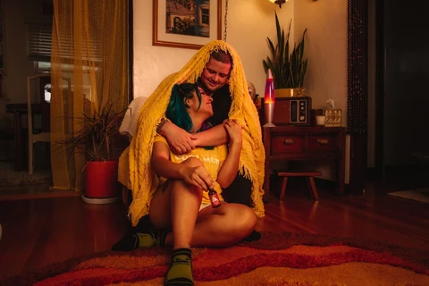 A man with a blanket over his head embraces his girlfriend on the floor of their living room