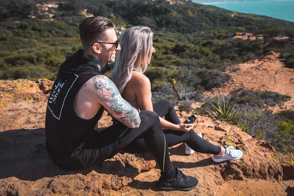 A male hiker embraces his girlfriend as they look out on the ocean from the top of a mountain