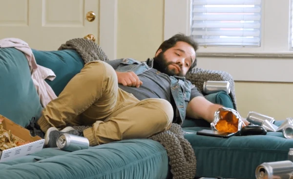 Man is hungover on the couch after a night of partying and his place is trashed
