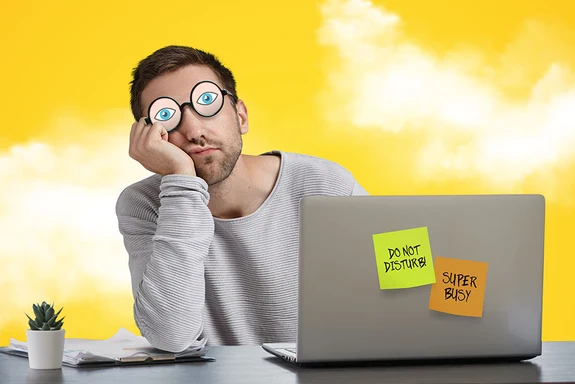 A man has fake glasses with painted eyes on them while he sleeps at work and pretends he's on his computer