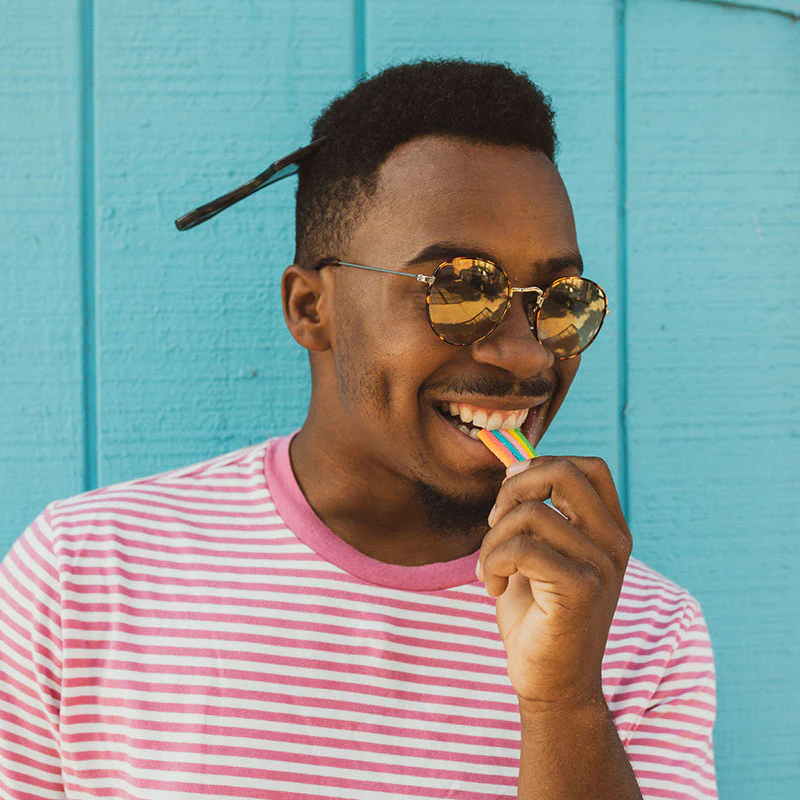 Man in sunglasses and striped shirt enjoying a rainbow-colored Sunday Scaries candy against a blue background.