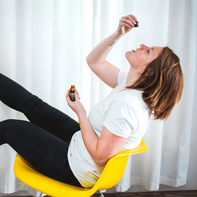 A woman tilts her head back on while sitting on a yellow chair and drops some CBD oil in her mouth